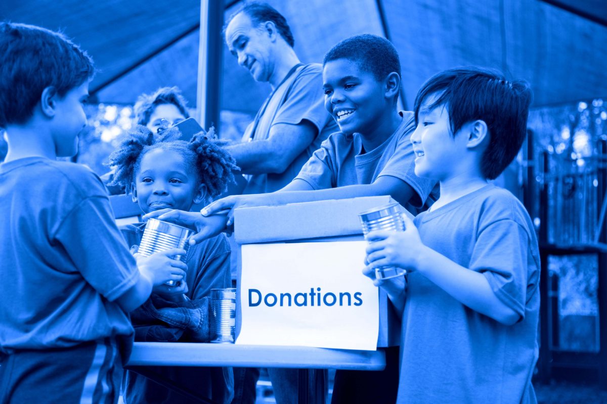 Multi-ethnic group of children on a soccer team work together to organize a charity drive for a local natural disaster event in their area. The children set up a table at their local park to gather canned goods and clothing for the victims affected by the disaster. Their team coach helps with the event.